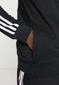 Svart träningsjacka med ribbad textur, vita randiga accenter på ärmarna och en sidoficka integrerad i designen.