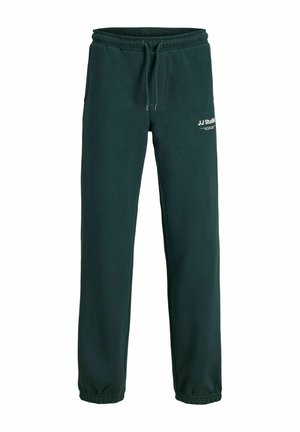 Garçon blond debout et souriant, portant un survêtement JJ Studios vert foncé avec un sweat à demi-zip et un pantalon de jogging assorti.