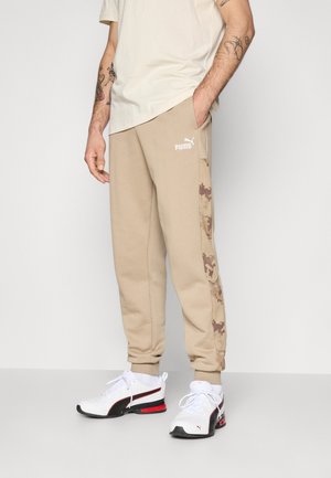 Man in beige Puma joggingbroek met camouflage zijstrepen en witte Puma sneakers, met een hand in de zak, staande tegen een effen achtergrond.