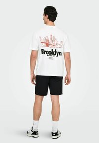 Hvid T-shirt med en rød skyline grafik og teksten "Brooklyn" på ryggen, parret med sorte shorts og sneakers.
