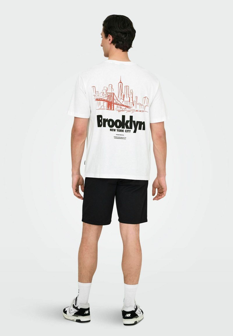 Hvid T-shirt med en rød skyline grafik og teksten "Brooklyn" på ryggen, parret med sorte shorts og sneakers.