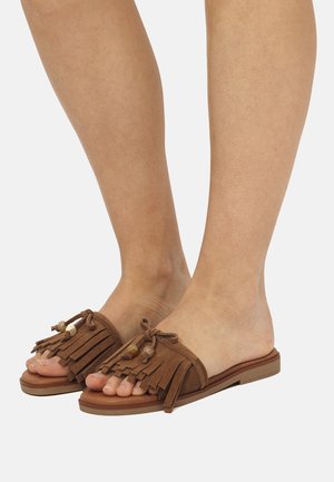 Pies con sandalias marrones sin cierre, con detalles de flecos y cuentas, sobre una suela plana, sobre un fondo blanco.