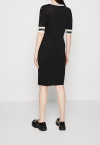 Femme portant une robe noire longueur genou avec des poignets rayés blancs et des manches en dentelle, associée à des mocassins noirs, debout devant un fond uni.