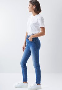 Vaqueros de tiro alto en azul con un corte ajustado, que cuentan con cinco bolsillos y cierre de botón. Combinados con una camiseta blanca lisa y zapatillas blancas.