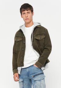 Jonge man in een wit T-shirt, grijze hoodie, olijfgroene knoopjack en lichtblauwe cargo jeans, poserend met één hand in zijn zak.