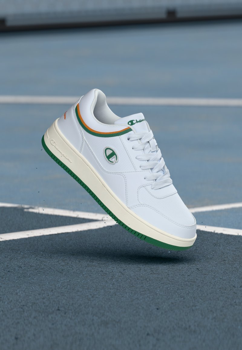 Baskets en cuir blanc avec des accents verts et orange, côtés texturés, lacets plats et semelle en caoutchouc. Présente un logo sur le côté.