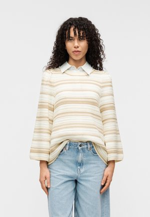 Pull rayé crème et beige, avec une texture côtelée, une coupe décontractée, et une chemise à col bleu clair en-dessous. Associé à un jean clair.