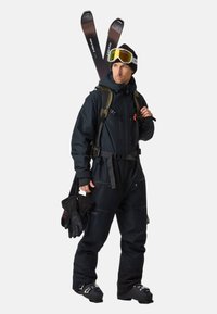 Zwarte skikleding met een ritssluiting jas, verstelbare straps en bijpassende broek. Voorzien van goggles, handschoenen en skis. Geweven, duurzaam materiaal.
