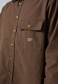 Smeđa pamučna košulja na kopčanje, s džepom na prsima, smeđim gumbima i bijelim izvezenim logotipom na džepu.