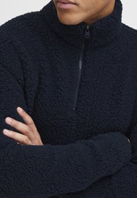 Marineblå fleecegenser med høy krage og kvart glidelås. Stoffet er teksturert og mykt, med en tykk, myk pels.