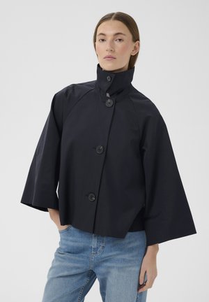 Kvinde iført en sort jakke med brede ærmer og knapper, høj krave og lyseblå jeans, stående med den ene hånd i lommen.