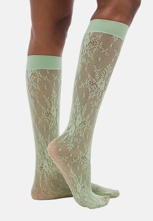 Ceļgalu augstuma zeķes gaiši zaļā mežģīņu materiālā ar ziedu rakstu un vienkrāsainu zaļu joslu augšpusē, ar caurspīdīgu, teksturētu dizainu.