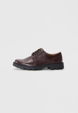 Zapato de piel marrón con cordones, punta redondeada, suela de goma negra y detalles en marrón contrastante en la lengüeta y el forro.