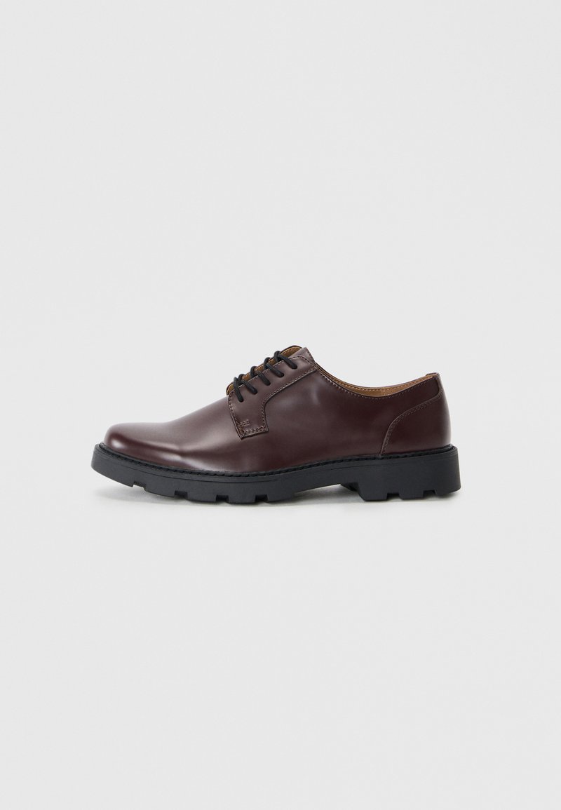 Zapato de piel marrón con cordones, punta redondeada, suela de goma negra y detalles en marrón contrastante en la lengüeta y el forro.