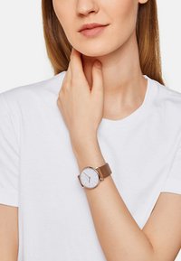 Montre-bracelet en or rose avec un cadran blanc minimaliste, un bracelet en cuir marron, un boîtier rond et des marqueurs d'heure simples, portée sur un poignet nu.