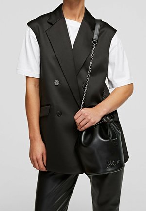 Person wearing a white t-shirt under a black sleeveless double-breasted blazer, holding a black leather drawstring bag with chain strap.