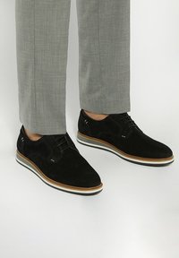 Chaussures habillées en cuir velours noir avec un bout arrondi, semelle blanche et marron, et un design à lacets bicolore. Le pantalon est gris clair avec un motif texturé.