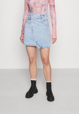 Persona vistiendo falda de mezclilla asimétrica azul claro, blusa transparente rosa de manga larga, calcetines beige y botas negras gruesas con cordones.