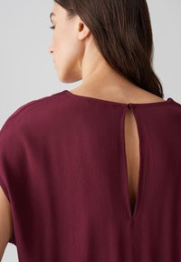 Bordeauxfarbenes Oberteil mit kurzen Ärmeln, das über ein Schlüsselrücken-Detail verfügt. Glatte Textur mit fließendem Design.