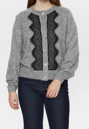 Femme portant un cardigan gris boutonné avec des volants en dentelle noire sur le devant, associé à un jean bleu foncé.