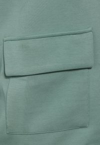 Textile vert clair avec une texture lisse. Présente une poche rectangulaire cousue sur la surface. Design simple et minimaliste.