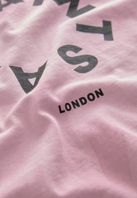 Rožinis medvilnės marškinėlis su dideliu juodu tekstu "LONDON" mažesniu juodu šriftu. Sklandaus paviršiaus, kasdieniško dizaino ir laisvo kirpimo.