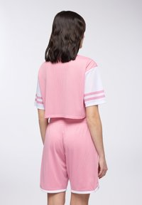 Różowy zestaw odzieży sportowej z siateczki, składający się z krótkiego topu z białymi rękawami i paskowanymi akcentami, w połączeniu z dopasowanymi szortami i białym wykończeniem.