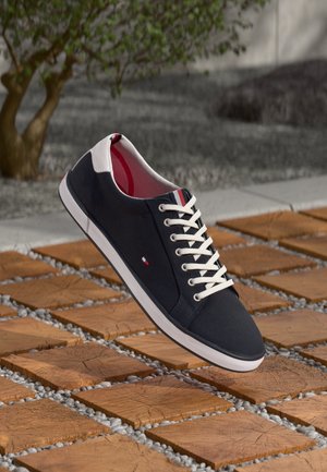 Marineblauer Sneaker mit weißen Schnürsenkeln und weißer Sohle, roten und weißen Akzenten, schwebend über hölzernen quadratischen Fliesen mit Kieseln im Außenbereich.