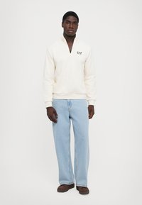 Hvit genser med halve glidelås, prydet med "EA7" logo. Kombinert med lyseblå brede jeans og brune slipsånskår.