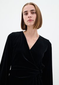 Svart sammetsklänning med långa ärmar, djup V-ringning och knytning i midjan. Den släta texturen och eleganta fall framhäver dess design.