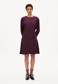 Kurzes lila Kleid mit langen Ärmeln, glatter Textur und lockerer Silhouette. Tragen mit rot gefleckten Socken und schwarzen Schuhen.