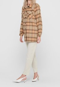 Veste-chemise à carreaux à motifs plaid dans des tons beige, brun et orange, avec de grandes poches poitrine, associée à un pantalon ajusté couleur crème et des talons blancs.