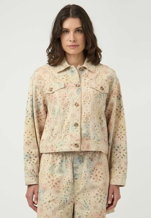 Femme portant une veste boutonnée beige à motif floral avec un pantalon taille haute assorti, debout devant un fond uni.