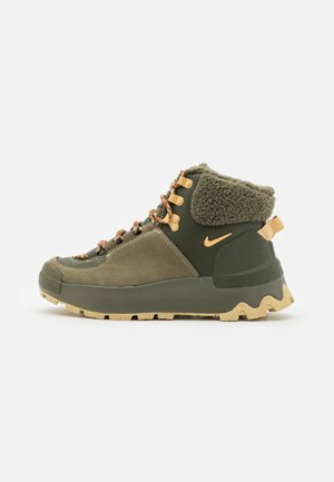 Bota de senderismo verde con una parte superior de ante texturizado y sintético, que presenta un collar de piel de fur, acentos naranjas y una suela robusta y con patrón.
