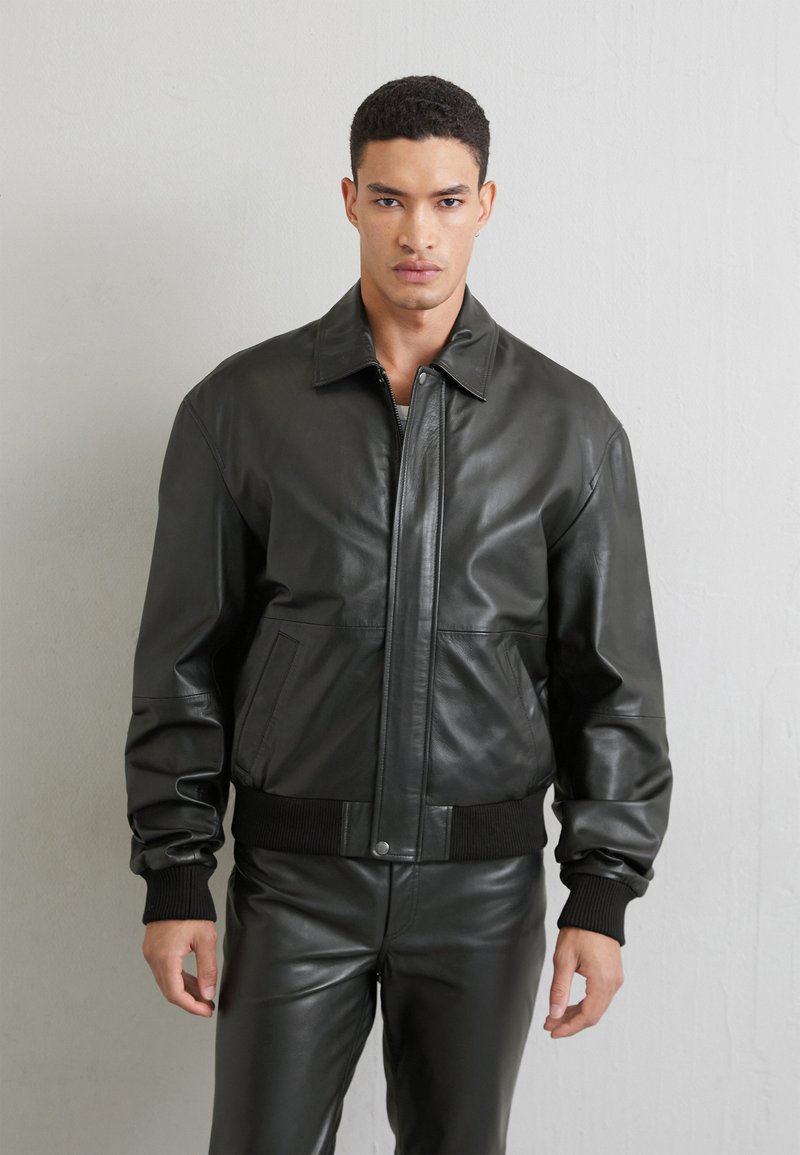 Svart läderbombarjacka med framdragkedja, två fickor med lappar och ribbade muddar. Slät textur och strukturerad design.