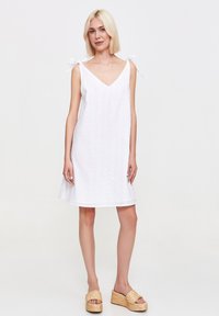 Modelka pozuje w białej sukience bez rękawów z kokardkami na ramionach, w połączeniu z słomianymi sandałami na platformie, na neutralnym tle.