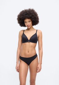 Γυναίκα με σγουρό αφρο χτένισμα, φορώντας μαύρο αθλητικό σουτιέν και ταιριαστό εσώρουχο, στέκεται μπροστά σε ένα απλό φωτεινό φόντο.