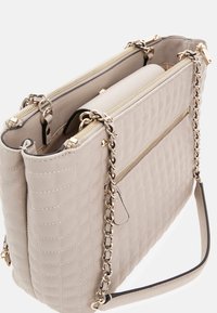 Bolso de mano acolchado beige con una superficie texturizada, cierres de doble cremallera, correa de cadena en tono dorado y bolsillo delantero con cremallera.