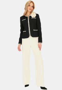 Czarna marynarka tweedowa z białymi wykończeniami, z dwoma przednimi kieszeniami i akcentem z tkaninowego kwiatu. Połączona z kremowymi spodniami w szerokim kroju.