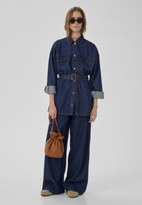 Žena oblečená v tmavě modré džínové bundě s páskem a širokých džínách, držící hnědou koženou tašku na stahovací šňůrku, s brýlemi na slunce a béžovými botami.