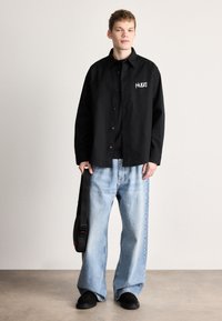 Μαύρο πουκάμισο με κουμπιά, άνετο ανοιχτό μπλε τζιν, μαύρα παπούτσια και μαύρη τσάντα με κόκκινες λεπτομέρειες, στέκεται μπροστά σε ουδέτερο φόντο.