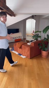 Caixa de cartão rotulada "Zalando" segurada por uma pessoa com uma camisa clara, em pé perto de um sofá de veludo cotelê laranja e uma planta verde em vaso.