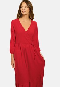 Vestido rojo ajustado con escote en V, mangas tres cuartos, detalle de lazo en la cintura y dobladillo en capas. Hecho de tela texturizada.