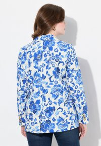 Kukallinen bleiseri valkoisena ja sinisenä, jossa on rakenteellinen muotoilu, pitkät hihat ja kultaiset napit hihansuissa.