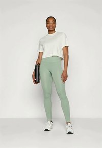 Zelené rebrované legíny v kombinácii s bielym skráteným tričkom, drží čiernu fľašu na vodu, s bielymi teniskami na neutrálnom pozadí.