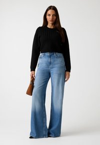 Maglione nero lavorato a maglia con motivo testurizzato, abbinato a jeans a gamba larga blu chiaro con una sfumatura graduale. Il modello tiene una piccola borsa marrone.