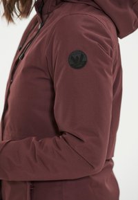 Zbliżenie osoby noszącej bordową kurtkę z kapturem, z czarną okrągłą naszywką na górnej części ramienia i czarnymi sznurkami przy kapturze.