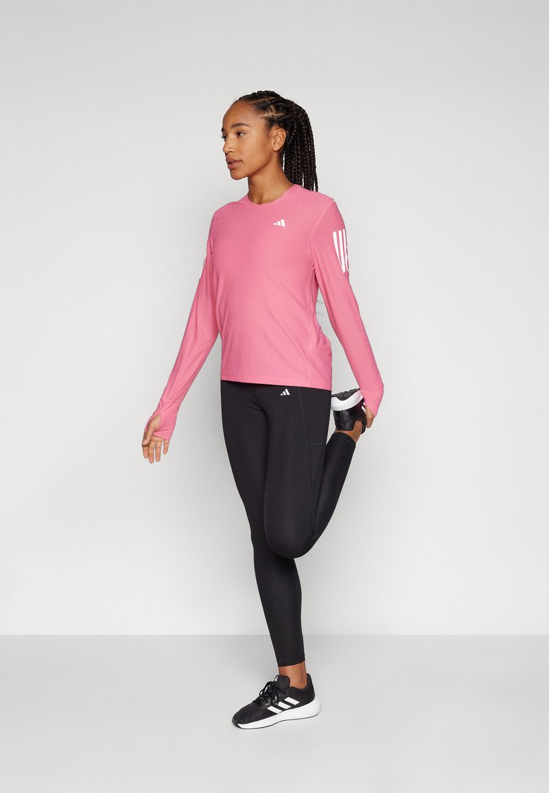Rosa langärmliges Sportoberteil mit weißem Logo, kombiniert mit schwarzen Leggings und schwarzen Sportschuhen. Die Person dehnt ein Bein nach hinten.