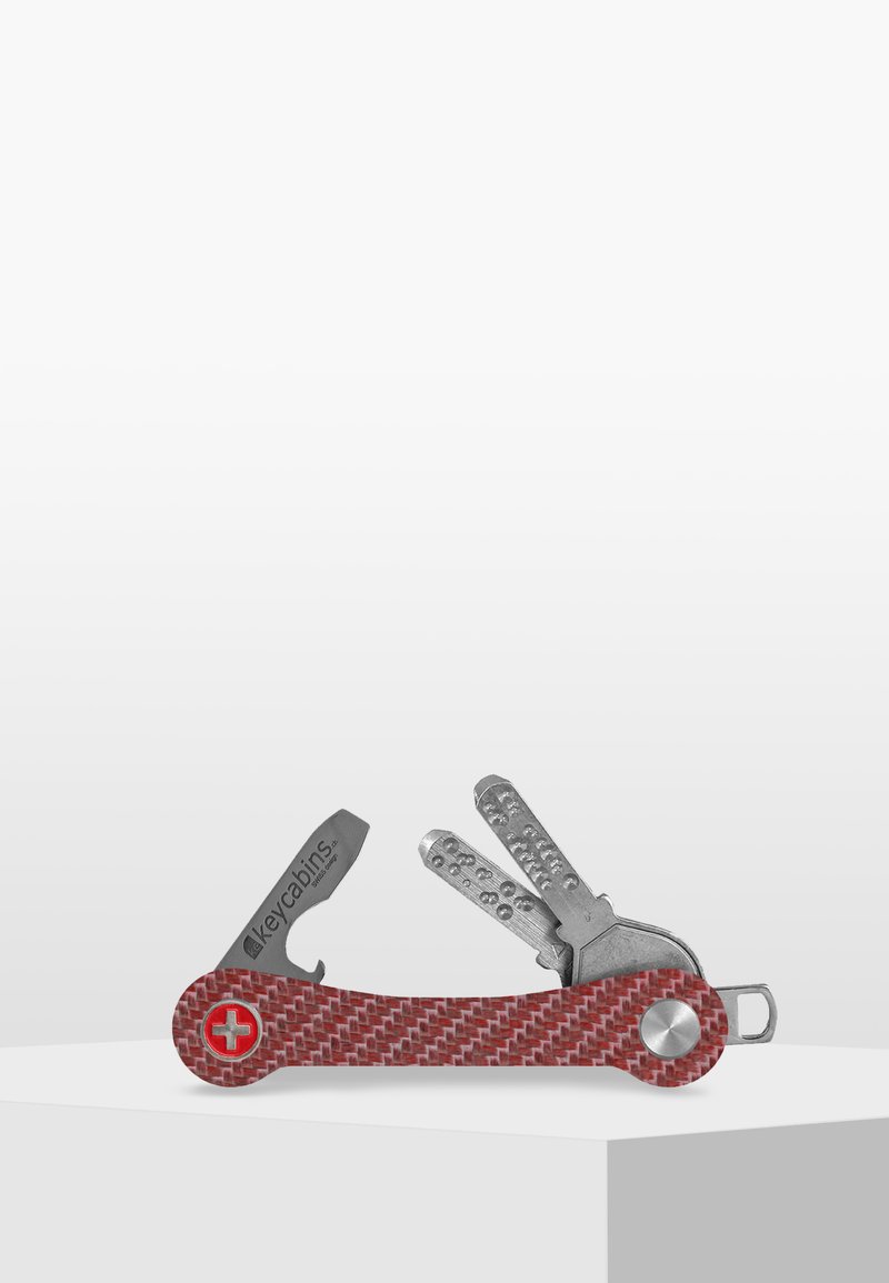 Röd mönstrad multifunktionstool med ett strukturerat handtag, som innehåller olika metallverktyg, inklusive en kniv och nyckelhållare, på en vit yta.