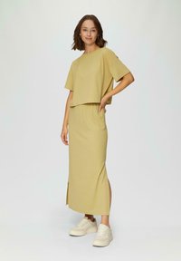 Gehaakt geel twee-delig outfit bestaande uit een kort mouwloze crop top en een lange rok met een zijsplit, gecombineerd met witte sneakers. Zachte textuur.